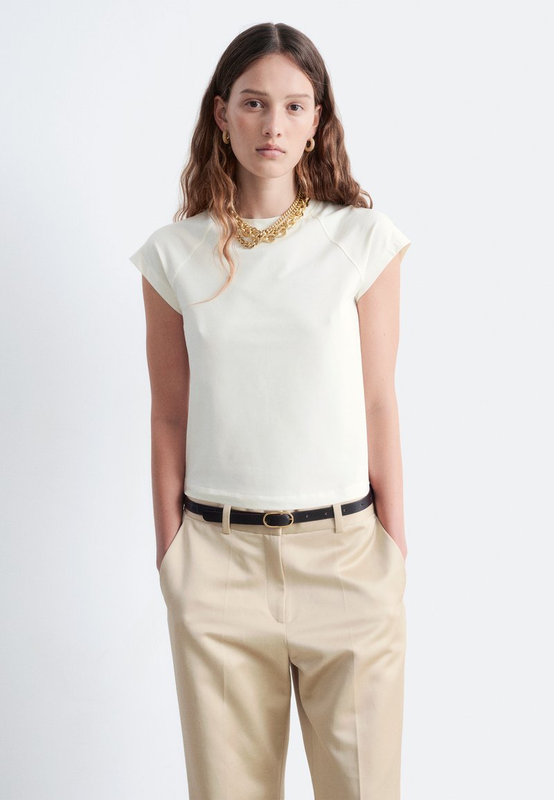 Weißes kurzärmliges Oberteil mit einer taillierten Passform, kombiniert mit beigen Hosen und einem schwarzen Gürtel. Das Model trägt eine geschichtete goldene Halskette.