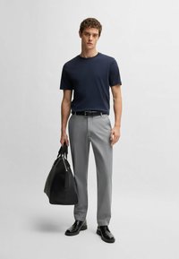 Navy t-shirt, light gray trousers, black belt, and black shoes. Holding a large black leather bag with double handles. Simple design.