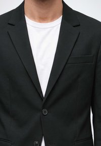 Homme portant un blazer noir sur un t-shirt blanc à col rond, photographié du cou jusqu'au milieu du torse sur un fond uni.