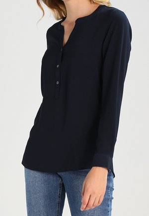 Woman wearing a long-sleeve navy blouse with a buttoned V-neck collar paired with blue jeans, standing against a plain background.