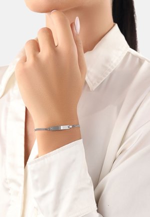 Bracciale d'argento con un ciondolo piatto e rettangolare su una catena strutturata, indossato su una mano su uno sfondo di camicia bianca.