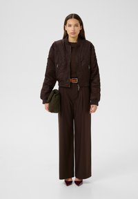 Femeie îmbrăcată cu o jachetă bomber maro închis, pantaloni largi asortați, curea maro, ținând o geantă verde măslin, stând pe un fundal simplu.