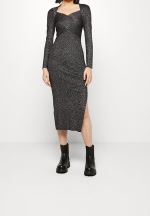 Woman wearing a long-sleeve, sparkly black midi dress with a side slit and black chunky ankle boots, standing against a plain white background.