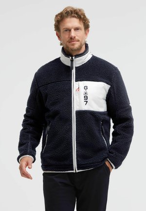Man wearing black and white fleece jacket with front zipper and "G97" logo on chest pocket, standing against a plain light background.