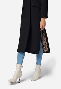 Schwarzer langer Mantel aus strukturiertem Stoff, mit seitlichem Schlitz, der ein Leopardenfutter zeigt, getragen über blauen Skinny Jeans und cremefarbenen Ankle-Booties.