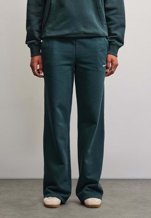 Person iført mørkegrønne løse joggingbukser og matchende hættetrøje, stående på gråt gulv med beige sneakers og sølvringe på fingrene.
