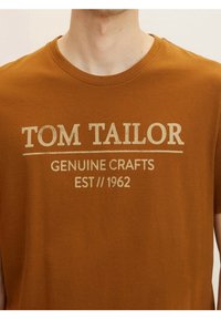 Hnědé bavlněné tričko s nápisem "TOM TAILOR" v tučném krémovém písmu, pod ním "GENUINE CRAFTS" a "EST // 1962". Krátké rukávy.
