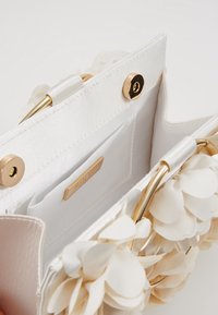 Sac à main blanc avec ornements floraux, ferrures dorées et texture satinée. Intérieur doté d'une poche plate et d'une étiquette de marque.