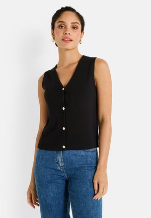 Femme portant un top sans manches noir à boutons et un jean bleu, se tenant contre un fond blanc uni.