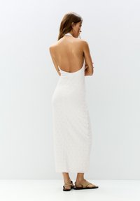 Bílá halter maxi šaty bez zad s květinovým výřezovým vzorem a texturovanou tkaninou. Nosené s béžovými sandály, které ukazují otevřený zadní design.