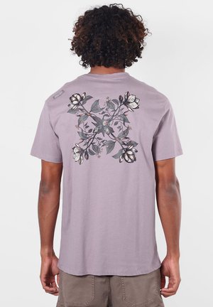Homme aux cheveux bouclés portant un t-shirt violet clair avec un motif floral symétrique dans le dos, associé à un pantalon beige.