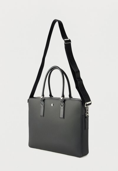 Serviette rectangulaire noire avec deux poignées et une bandoulière réglable, présentant un embossage du logo Lacoste.
