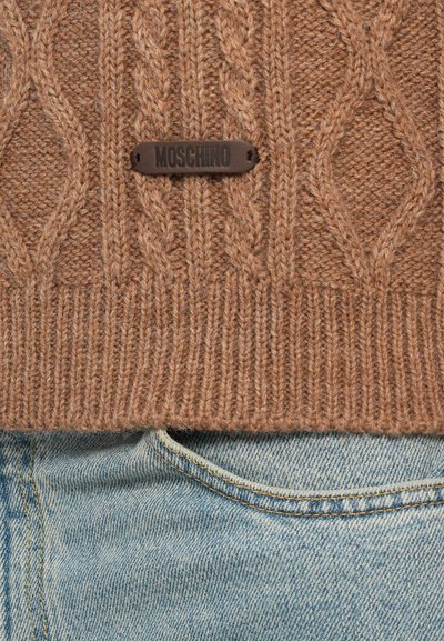 Brun stickad tröja med kabelmönster och en svart Moschino-logotagg, tillsammans med ljusblå jeans. Texturerad tygdetalj synlig.