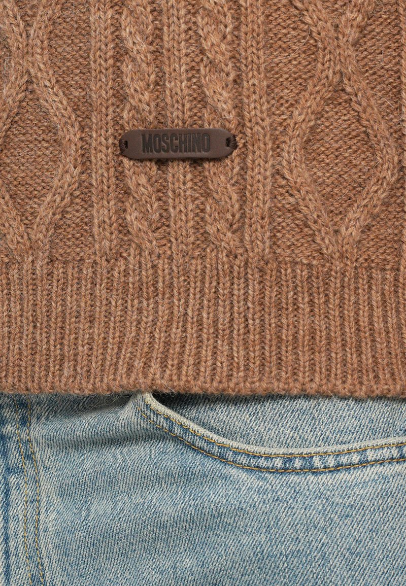 Bruine gebreide trui met een kabelpatroon en een zwart Moschino logo-label, gecombineerd met lichtblauwe denim jeans. Textuurdetail van de stof is zichtbaar.