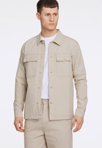 Ljust beige knäppjacka med lång ärm, två bröstfickor, mjuk tygtextur, kombinerad med en vit T-shirt och matchande beige byxor.