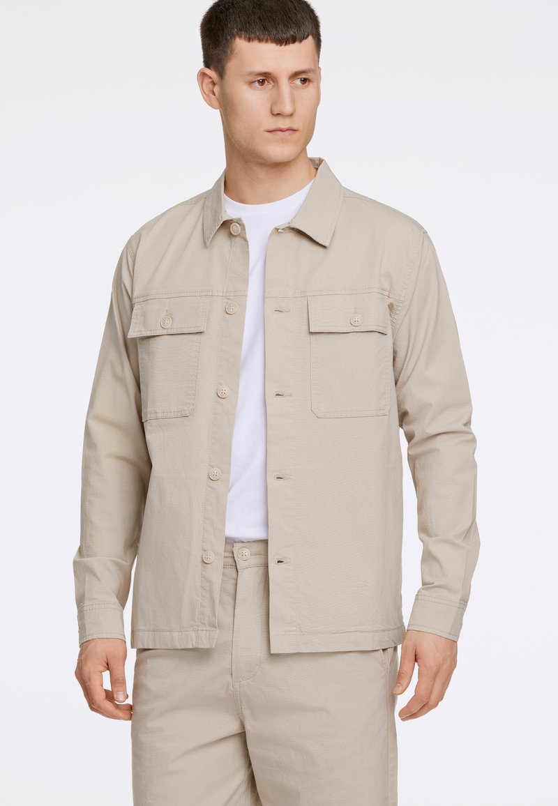 Ljust beige knäppjacka med lång ärm, två bröstfickor, mjuk tygtextur, kombinerad med en vit T-shirt och matchande beige byxor.