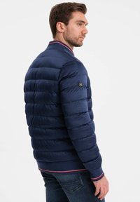 Marineblauer Daunenjacke mit horizontalen Nähten, geripptem Kragen und Bündchen mit roten und weißen Akzenten. Mit Reißverschlusstaschen an den Seiten.