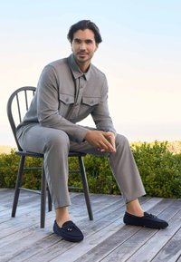 Graue, knopfbare Jacke mit zwei Brusttaschen und passender Hose; marineblaue Slippers mit Schnürdetails; sitzt auf einem schwarzen Holzstuhl.