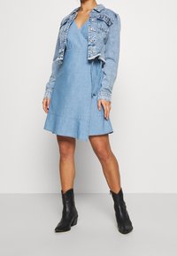 Dżinsowa kurtka ma skrócony krój z postrzępionym dołem, nałożona na jasnoniebieską sukienkę kopertową z zwiewnym dołem. Czarny botki za kostkę dopełniają całość.