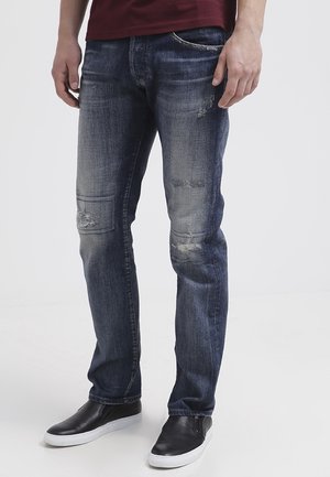 Mann trägt verblasste, abgenutzte blaue Jeans mit kleinen Rissen, schwarze Slipper mit weißen Sohlen und ein dunkelrotes Hemd, das am Bund sichtbar ist.