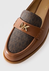 Bruine leren instapschoenen met een gestructureerd donkerbruin voorpaneel en een gouden "MK"-logo op de band, gestikte details en een rubberen zool.