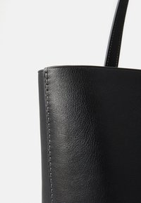 Geantă tote din piele neagră, cu o textură netedă, o formă structurată și detalii de cosătură vizibile pe latura acesteia. Design simplu al mânerei.