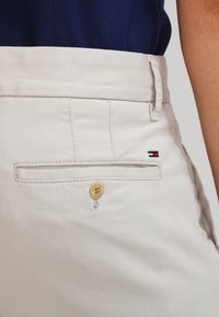 Světlé béžové bavlněné kalhoty s kapsou na zadní straně se zapínáním na knoflík a vyšitými akcenty v červené, bílé a modré barvě. Hladká textura, upravený střih.