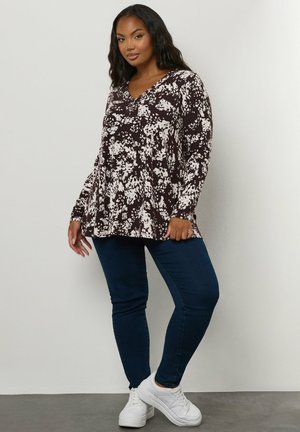 Longsleeve-Oberteil in Dunkelbraun mit einem weißen Blumenmuster, mit einem V-Ausschnitt und einem ausgestellten Saum. Dazu kombiniert werden Dunkelblaue Jeans und weiße Sneakers.