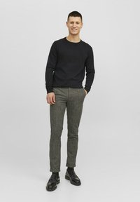 Pull noir, pantalon gris à carreaux slim-fit et mocassins noirs. Le mannequin est debout devant un fond uni, regardant droit devant lui.