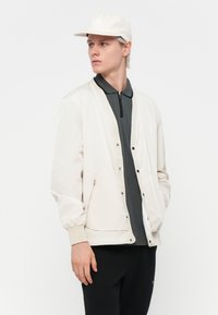 Light beige bomber jacket with elastic cuffs and snap buttons, layered over a dark polo shirt, complemented by a matching cap. Black pants.
