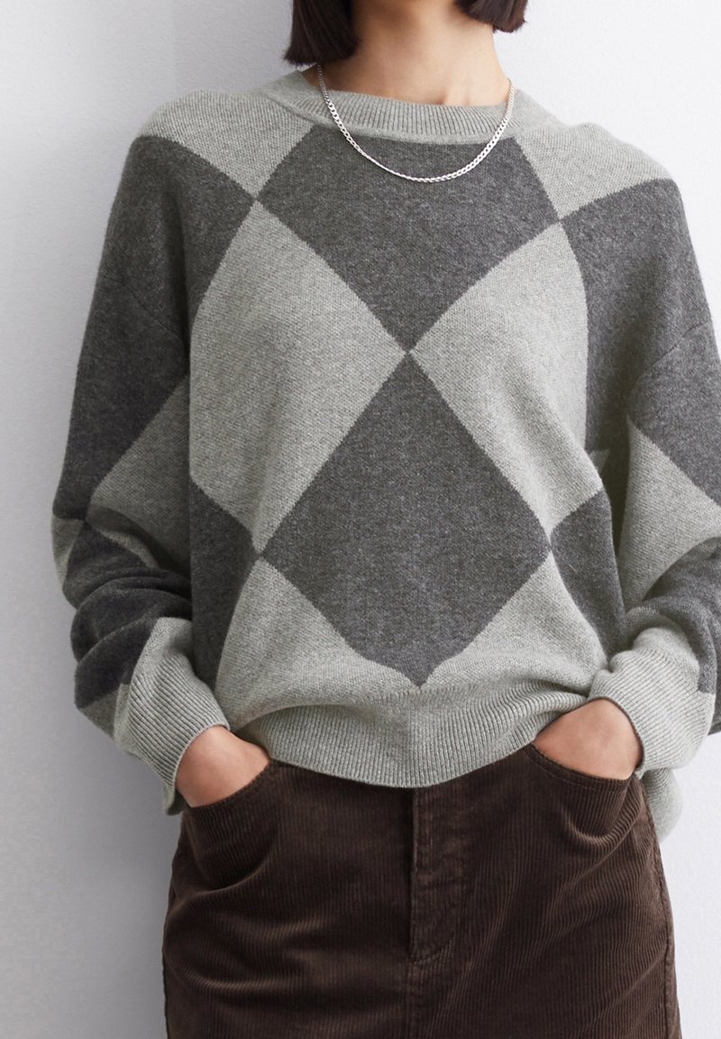 Femme portant un pull gris à motif losanges, un collier chaîne argenté et un pantalon en velours côtelé marron, les mains dans les poches, contre un mur blanc.