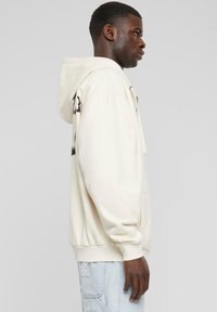 Hooded sweatshirt in light cream, featuring a relaxed fit, ribbed cuffs, and graphic detailing on the sleeves. Smooth texture.