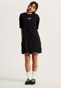 Robe t-shirt en coton noir à manches courtes, arborant un logo Vans sur la poitrine. Associée à des chaussettes blanches et des chaussures noires décorées de pois.