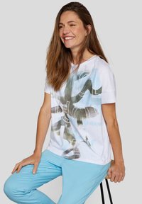 Weißes Grafik-T-Shirt mit einem blauen und grünen abstrakten Design. Kombiniert mit hellblauen Hosen; der Stoff wirkt weich und leicht.