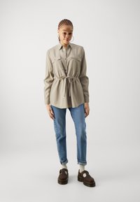 Light gray, long-sleeve shirt with two chest pockets and a drawstring waist, paired with blue jeans and brown clogs. Bright background.