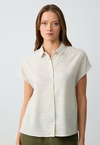 Camicia corta beige chiaro realizzata in tessuto testurizzato con colletto e chiusura a bottone, caratterizzata da sottili strisce orizzontali.