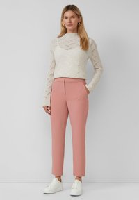 Vrouw in een beige kanten blouse met lange mouwen, stofroze broek, witte sneakers en gouden oorbellen op een effen grijze achtergrond.