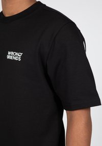 Černé bavlněné tričko s krátkým rukávem a kulatým výstřihem, na hrudi s bílým logem "WRONG FRIENDS". Hladká textura.