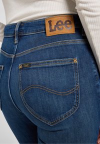 Pantalones vaqueros de mezclilla azul con un parche de cuero con el logo de Lee en la cintura, costuras amarillas y un diseño de bolsillo curvado.