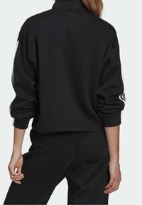 Svart kort sweatshirt med hög krage, ribbade ärmslut och tre vita ränder längs ärmarna. Materialet verkar mjukt och texturerat.