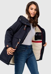 Marineblaue Jacke mit sandfarbenen Akzenten und einem plüschigen beigen Innenfutter; verfügt über einen Reißverschluss vorne und Innentaschen für die Aufbewahrung.