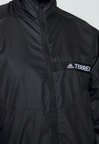 Chaqueta negra Adidas Terrex con cremallera frontal y cuello alto, centrando la atención en el logo en la zona del pecho.