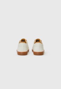 White leather shoes with a rounded toe, tan accents on the back, and rubber sole. Rear view emphasizes sleek design and contrasting colors.