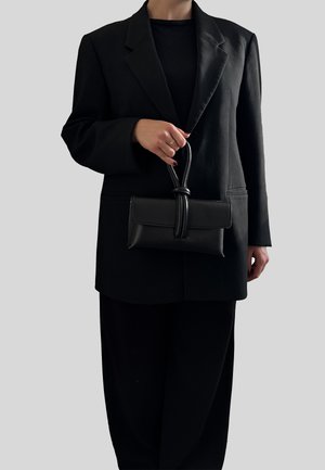 Borsa a mano nera in pelle con manico annodato, forma rettangolare, superficie testurizzata, tenuta davanti a una persona che indossa un blazer e un top neri.