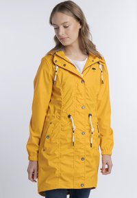 Egy hosszú, sárga esőkabátot viselő nő, felhúzott kapucnival, fehér húzózsinórokkal és gombos részletekkel, lefelé néz egy sima fehér háttér előtt.