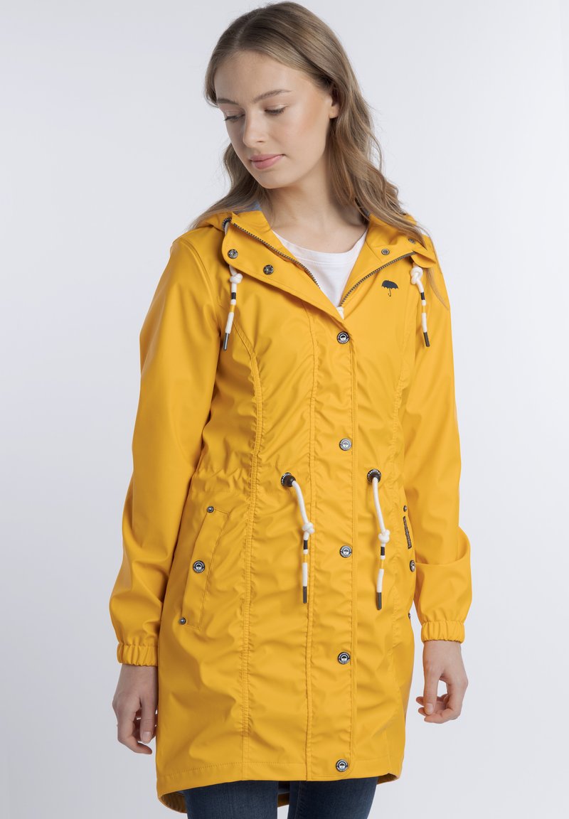 Egy hosszú, sárga esőkabátot viselő nő, felhúzott kapucnival, fehér húzózsinórokkal és gombos részletekkel, lefelé néz egy sima fehér háttér előtt.