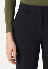 Pantalons ajustés bleu marine foncé avec des rayures fines. Présentent un devant plat, des poches latérales et une texture lisse. Un haut olive à manches longues est visible.
