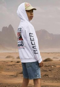 Witte hoodie met graphics op de voorkant en mouwen, gecombineerd met blauwe denim shorts. Een tan cap maakt de outfit compleet. Achtergrond toont een woestijnlandschap.