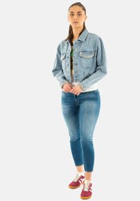 Chaqueta de mezclilla azul claro con bolsillos adornados, llevada sobre una camiseta blanca y combinada con jeans ajustados azules y zapatillas burdeos.
