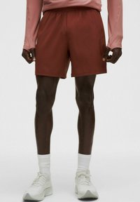 Bărbat îmbrăcat în pantaloni scurți atletici de culoare ruginită, tricou cu mânecă lungă roz, șosete albe până la glezne și pantofi de alergare albi, pe un fundal simplu.